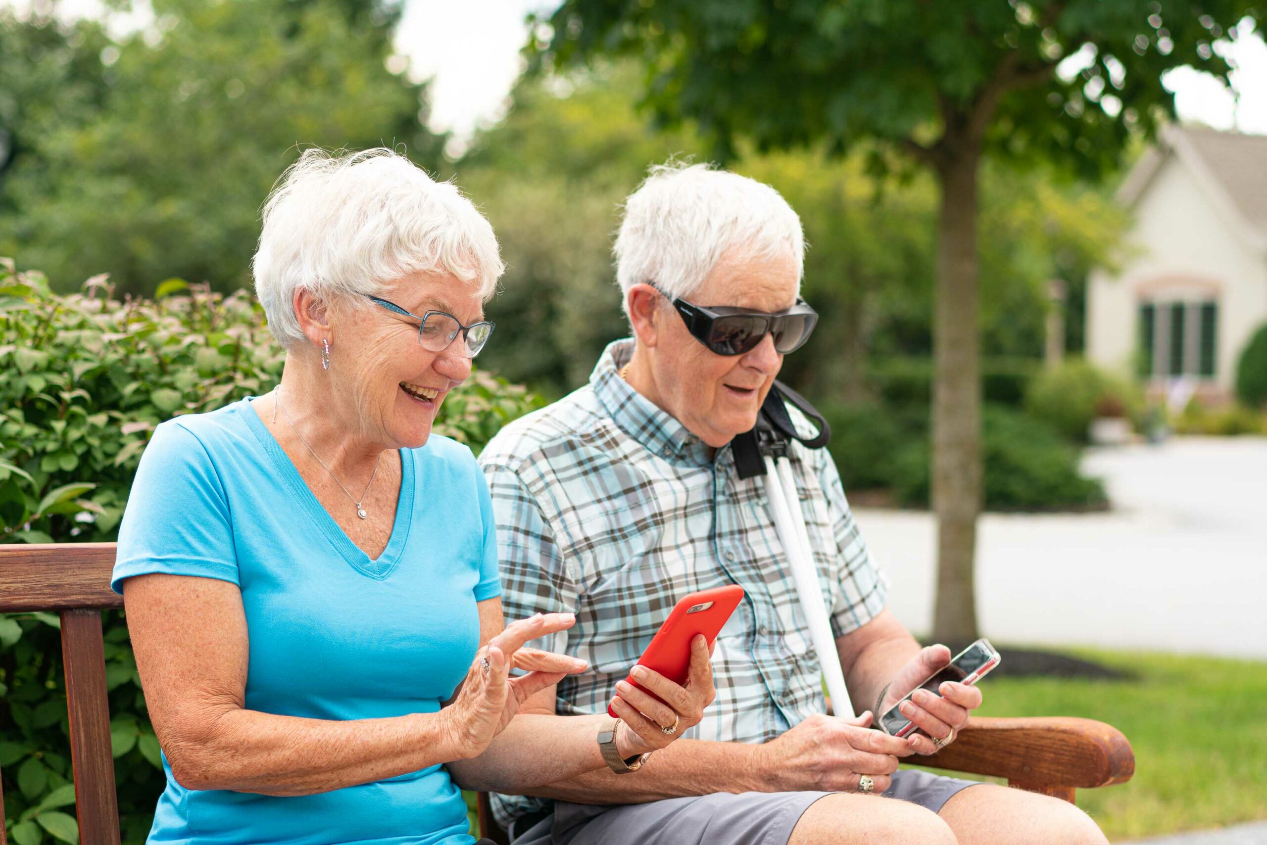 A man and woman sit on a bench together. The woman, with silver hair and blue shirt, is looking at her cell phone. The man, with silver hair and plaid shirt, wearing sun shields, is also looking at a cell phone.