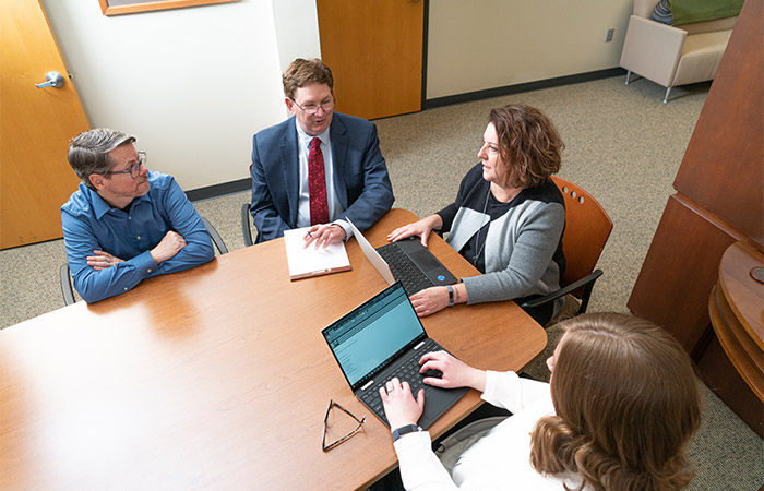 Two men and two women surround a conference table while typing on computers and writing on a note pad.