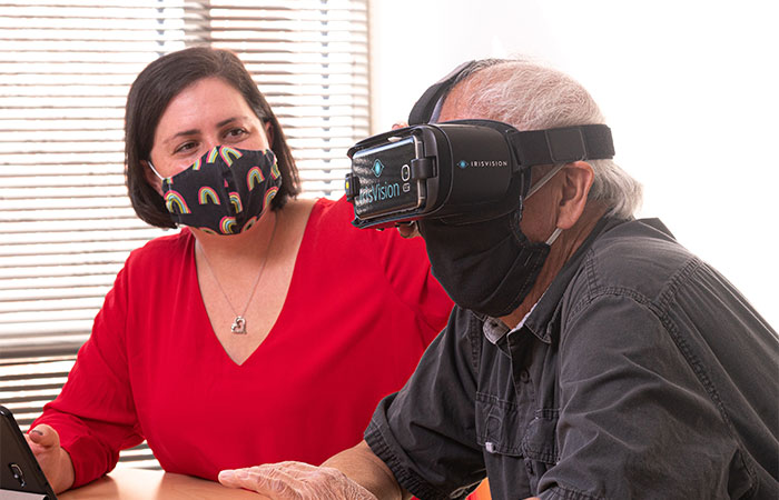 A middle aged woman sits beside a middle aged man. He is wearing Iris VIsion smart glasses and the woman helps to adjust them on his face