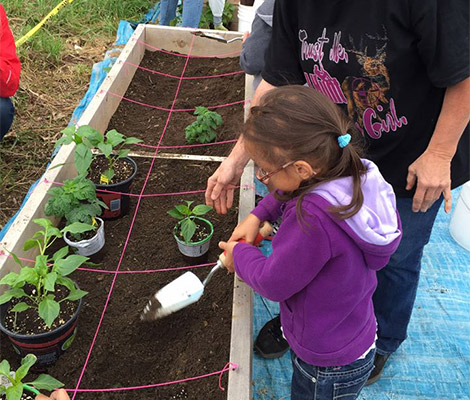 A little girl holds a small garden shovel to dirt in a raised garden bed, while a middle aged female volunteer looks on.