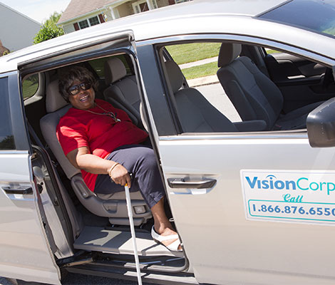 An older woman in a bright red shirt sits in the back seat of a VisionCorps van, waiting for the volunteer driver to transport her.